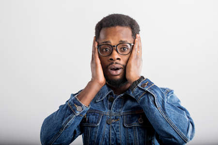 Portrait of surprised dark skinned man in glasses touching his face and opening mouth, being in shok, astonished with some unexpected news isolated on gray background. Human facial expressions.の写真素材