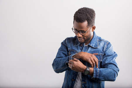 Young afro american unshaven man in glasses, with wristwatches, fastens button on sleeve cuffs of denim jacket smiling looking down isolated on gray background with copy space.の写真素材