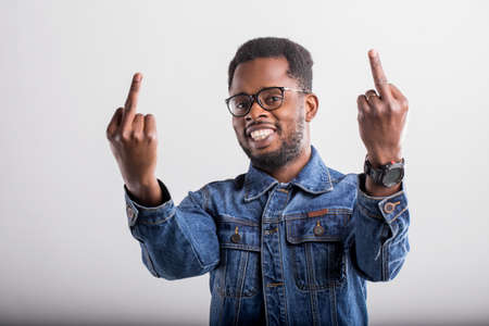 Portrait of cheerful carefree young african man with curly short hair dressed in trendy denim clothes, showing middle fingers and smiling broadly with white teeth over gray studio wall.の写真素材