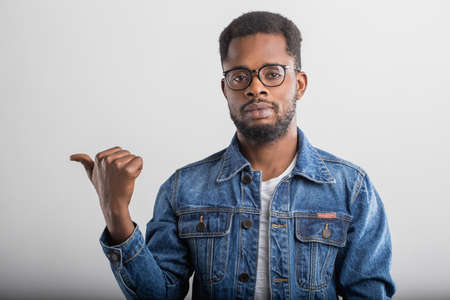Waist-up shot of confident carefree african man with small beard in glasses wearing denim jacket pointing left with thumb and looking at camera standing over gray background.の写真素材