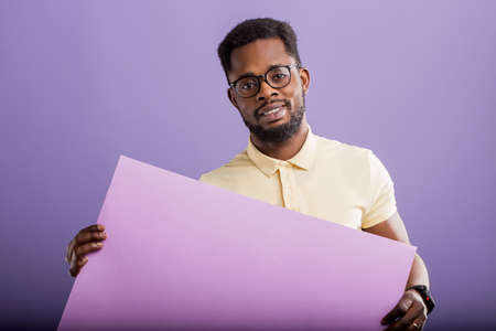 Picture of young african american man holding lilac blank board looking at camera on purple background, copy space. Handsome serious guy with blank placard ready for your text or product.の写真素材
