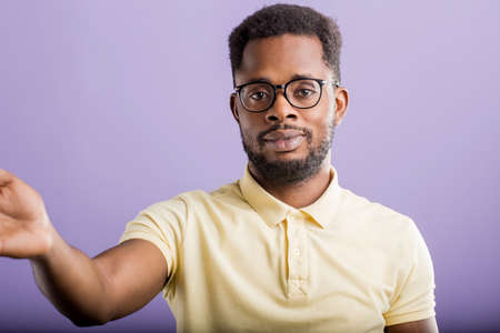 Closeup portrait of charming friendly african man in glasses pulling hand towards camera, smiling reaching out to take or give something, taking selfie on smartphone over purple background.の写真素材