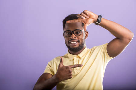 Studio portrait of happy carefree joyfull african american male model making frame gesture with fingers in front of his face, looking at camera, smiling broadly on lilac background.の写真素材