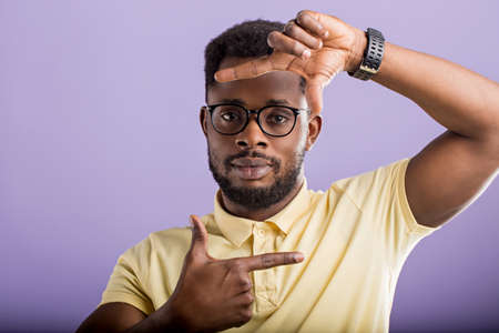 Studio portrait of intelligent confident handsome unshaven african man in glasses, wrist watch, making frame gesture with fingers in front of his face, looking at camera, isolated purple background.の写真素材
