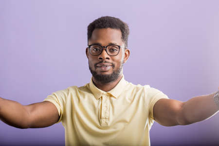 Friendly joyful handsome unshaven african american man wearing eyewear, yellow polo tshirt smiling and pulling hands towards camera wanting to hug, welcoming friends on lilac background.の写真素材