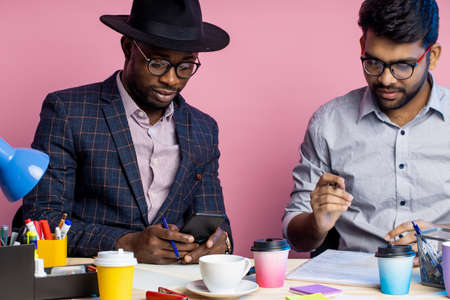 african american businessman and indian enterpreneur discussing business plans, sitting at table in office, wearing formal wear, smiling and laughing. Business, coworkers concept.の写真素材