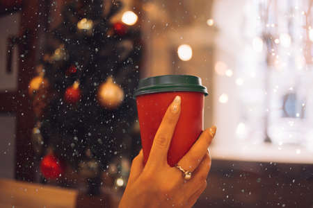 woman holding red paper cup of coffee or hot tea sitting in front of window with garland, christmas lights at cafe. New year, drinks, beverage concept.の写真素材