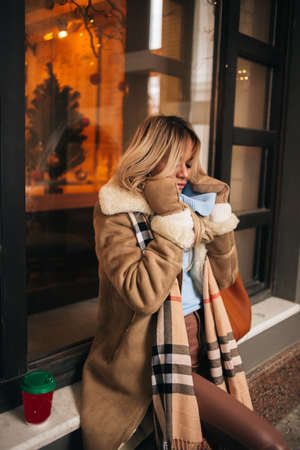 Beautiful blonde girl feeling cold, trying to protect herself from the freeze in city on winter day. Young caucasian woman waiting for friend on the street. Cold weather concept.の写真素材