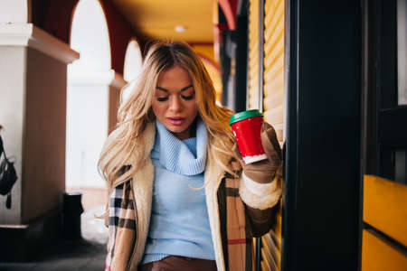 blonde girl walking around the city, smiling, drinking hot coffee, wearing stylish beige coat, mittens and scarf. Lifestyle, people, drinks concept.の写真素材