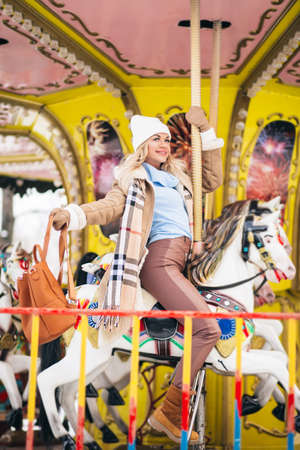Portrait of attractive lady in trendy clothes with big brown backpack riding merry-go-round holding cup of hot drink, dreaming, spending time in amusement park in weekend. Magic warm new year photo.の写真素材