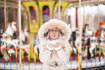 Joyful charming blonde young woman having fun in amusement park in winter. Closeup fashion portrait of happy girl blowing snowflakes from her hands. Enjoying nature.の写真素材