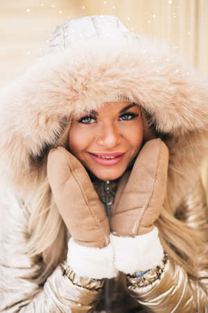 Outdoor portrait of young beautiful happy smiling girl posing on street, sitting on bench, looking at camera, touching cheeks with hands. Lady wearing stylish winter jumpsuit, boots. Female fashion.の写真素材