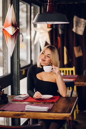 Smiling business woman in a restaurant holding a Cup of tea and looking at the camera, sitting near the window in a black dressの写真素材