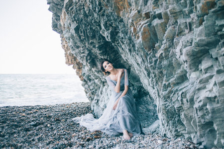 Beautiful brunette girl in blue dress sitting near rocks, sea, smiling, looking upの写真素材