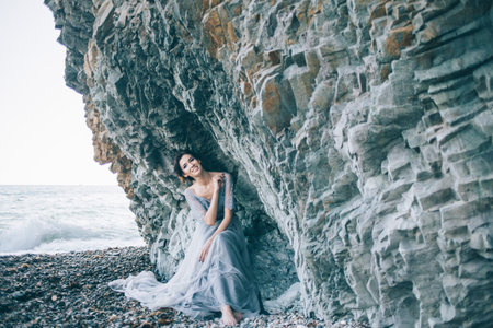 Beautiful brunette girl in blue dress sitting near the cliff of the sea, laughing, smilingの写真素材