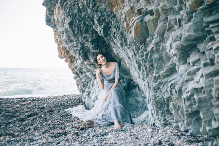 Beautiful brunette girl in blue dress sitting near the cliff of the sea, laughing, smilingの写真素材