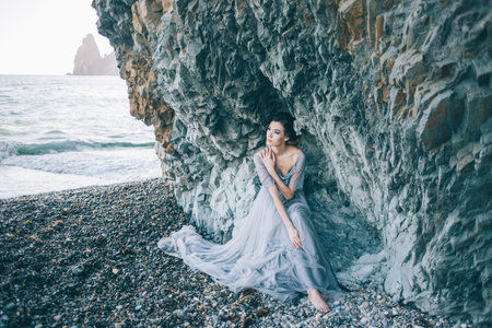 Beautiful brunette girl in a long gray dress with a veil sitting by the sea, near the cliff, looking profileの写真素材