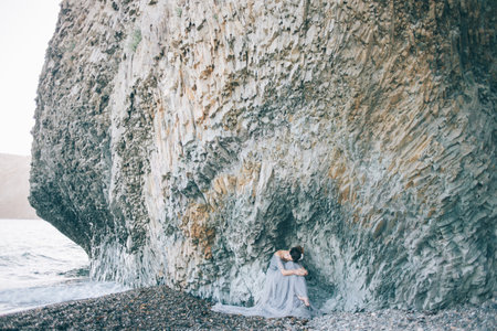 Beautiful girl bride in a long blue dress sitting on the sea near the cliff with his head bowed, hugging her kneesの写真素材