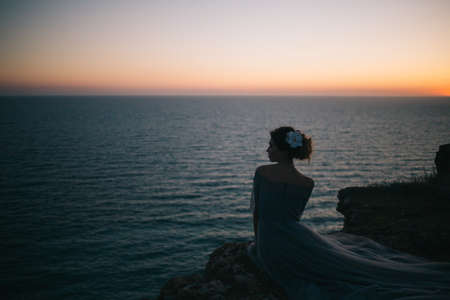 Beautiful girl in a long dress with a veil sitting on the beach at sunset, profileの写真素材
