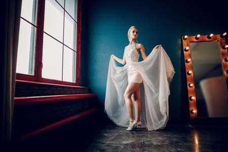 A slender girl in a light dress stands between the window and the mirrorの写真素材