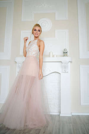 Smiling tall blonde in a light dress with tanned legs hidden by fabric looks to the side against the background of a beige wall with white stucco molding and a fireplace with candlesの写真素材