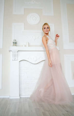 Playful bride with blond hair smiles and walks to the side against the background of a beige wall and a fake fireplace with candlesの写真素材