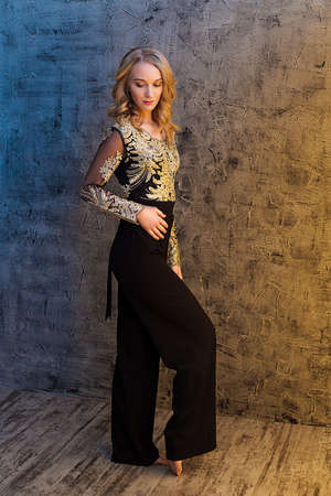 A curly-haired blonde girl stands in a trouser and a jacket with a gold pattern and looks at the floor. A model in a black and gold suit against a neutral wall with yellow and blue lighting.の写真素材