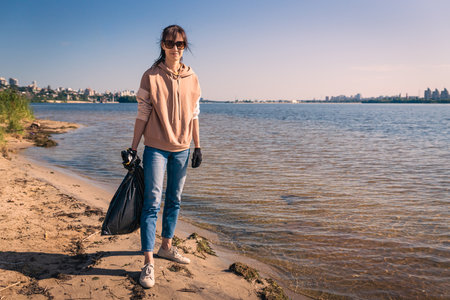Young woman with trash bag in beach cleans up plastic bottles