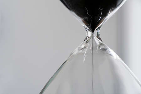 Black sand running through the bulbs of hourglass measuring the passing time in a countdown to a business deadline, urgency and running out of time on a light background with copy space close upの写真素材