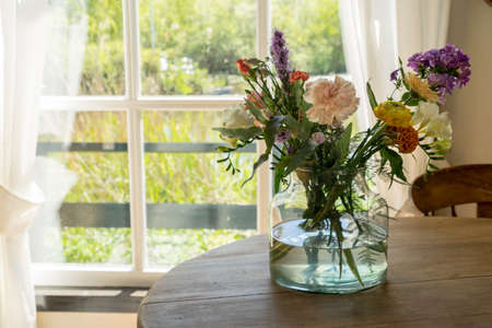 Vase with a mixed bouquet of flowers standing on a wooden table behind a window in the kitchenの写真素材