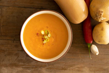 A view of bowl with delicious pumpkin soup, ingredients like pumpkin, potato, garlic and pepper on a wooden tableの写真素材