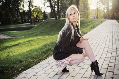 Young woman squats on sidewalk in city park in the autumn. The scene is shined behind by the sun の写真素材
