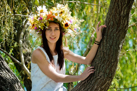 Beautiful brunette girl wreath of flowers is standing by a tree.の写真素材
