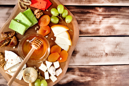 Top view of cheese plate - various types of cheese, honey, grapes, dried apricots, nuts and figs on a wooden board on dark wooden background.の写真素材