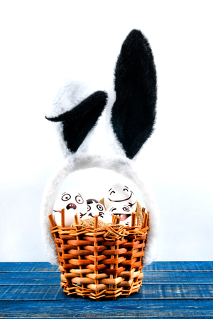 Funny easter eggs with different funny faces in the basket with rabbit ears  on white and blue background.の写真素材