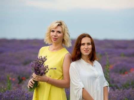 Two young girls with different hair color in white and yellow dresses, posing together in a lavender field.の写真素材