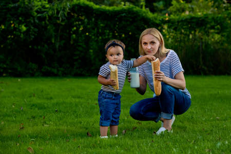 A young woman in jeans and a striped T-shirt gives her daughter fresh baguette. Family picnic.の写真素材