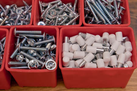 Various size and shape of screws, bolts, wooden pegs selection in red plastic tray box on a wooden...