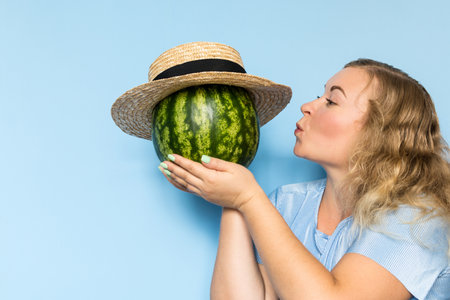 Young blonde woman holding and giving a kiss to watermelon in straw hat. Light blue background.の写真素材