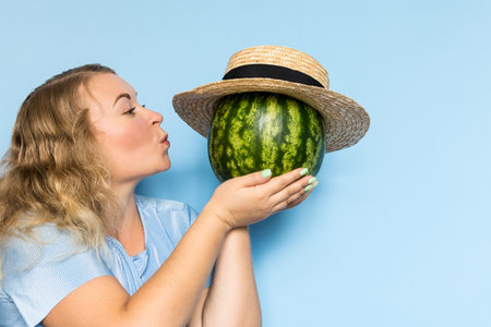 Young blonde woman holding and giving a kiss to watermelon in straw hat. Light blue background.の写真素材