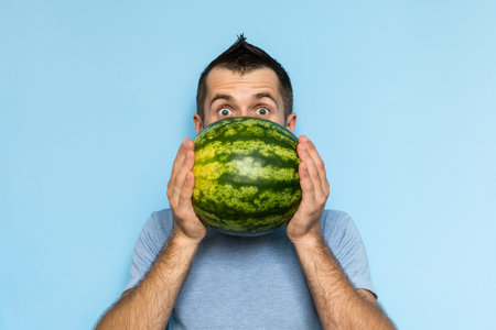 Young man holding a stripy watermelon in front of his face. Light blue background.の写真素材