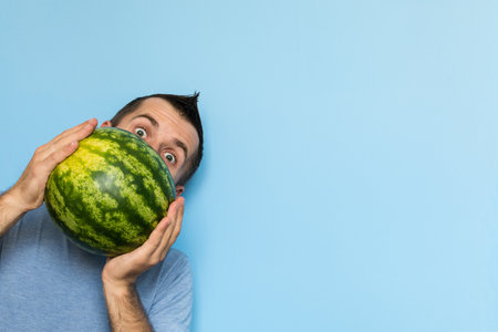 Young man holding a stripy watermelon in front of his face. Light blue background.の写真素材