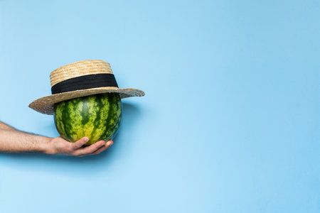 Man's hand holding a watermelon covered with straw hat. Light blue background.の写真素材