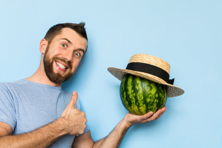 Young man holding a stripy watermelon covered with straw hat in front of his face. Light blue background, man showing thumb up.の写真素材