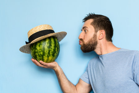 Young man holding and giving a kiss to a stripy watermelon in straw hat. Light blue background.の写真素材