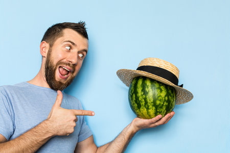 Young man holding a stripy watermelon covered with straw hat in front of his face. Light blue background, man showing hand shoot gun sign.の写真素材