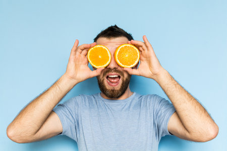 Young bearded and smiling man holding slices of orange in front of his eyes. Light blue background.の写真素材