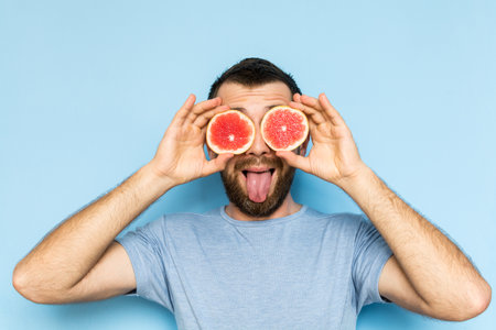 Young bearded man holding slices of grapefruit in front of his eyes. Light blue background, man putting out his tongue.の写真素材