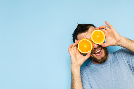 Young bearded and smiling man holding slices of orange in front of his eyes. Light blue background.の写真素材