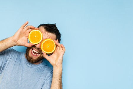 Young bearded and smiling man holding slices of orange in front of his eyes. Light blue background.の写真素材